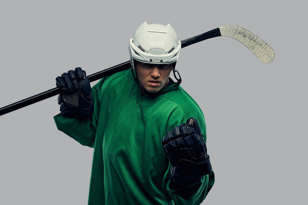 Angry professional hockey player in green sportswear standing with a hockey stick. Isolated on a gray background.  - Photo, Image