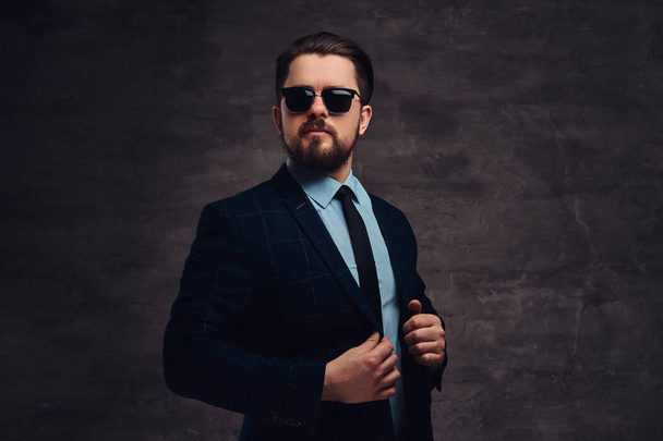 Confident handsome fashionable middle-aged man with beard and hairstyle dressed in an elegant formal suit and sunglasses on a textured dark background in studio. - Fotografie, Obrázek