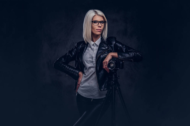 Blonde female photographer in trendy clothes posing while leaning on a professional camera with a tripod at a studio, looks at the camera. Isolated on a dark background. - Photo, Image