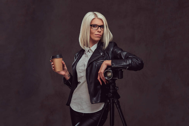 Muotokuva fiksu blondi naisvalokuvaaja trendikkäissä vaatteissa ja lasit pitää takeaway kahvia ja poseeraa samalla nojaten ammatillinen kamera jalusta studiossa, katsoo kameraa
 - Valokuva, kuva