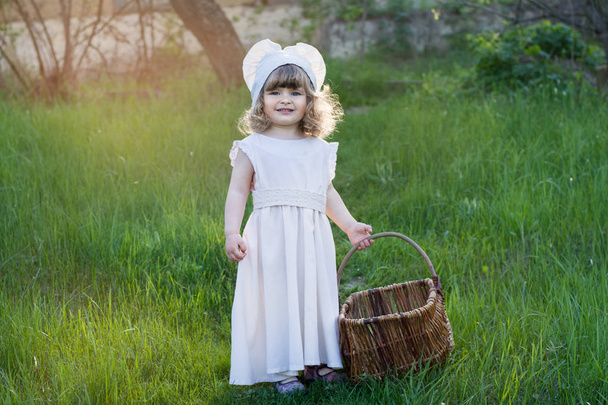 A beautiful little girl is walking in the countryside. A child in a white dress with a wicker basket in her hands. Sunset photo - Zdjęcie, obraz