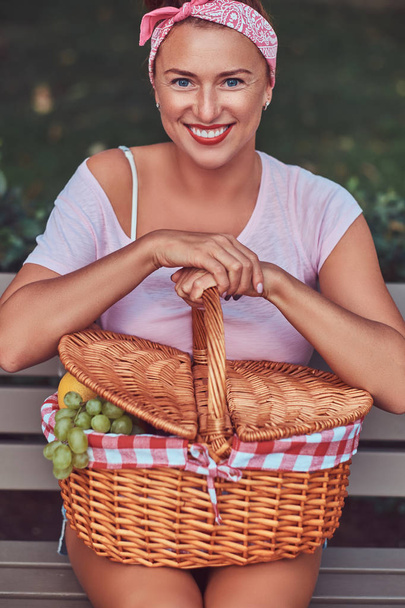 Happy beautiful redhead female wearing casual clothes holds a picnic basket while sitting on a bench in the park. - Zdjęcie, obraz