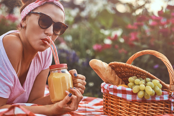 Onnellinen punapää keski-ikäinen nainen rento vaatteita otsanauha ja aurinkolasit nauttia piknikillä ulkona makaamassa huopa puistossa
. - Valokuva, kuva