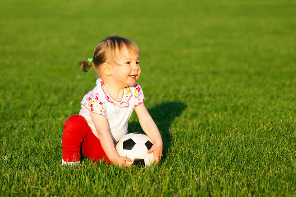 The smiling little cute baby girl is playing with small soccer ball for children on green grass playing field. - Photo, Image