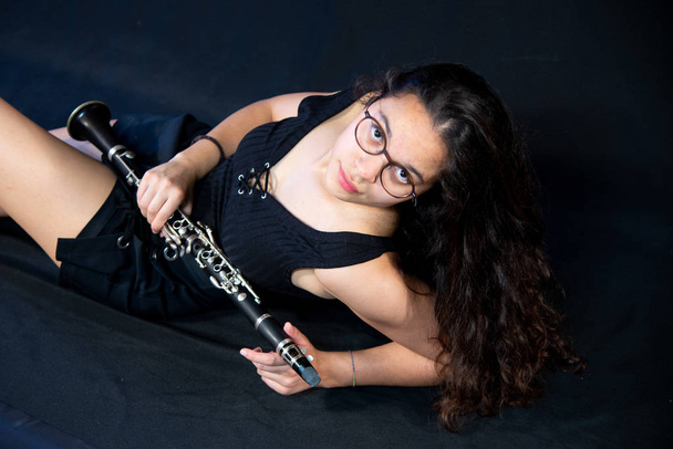 A brunette girl, with glasses, lying laughing and holding her clarinet music instrument isolated on a black background - Photo, Image