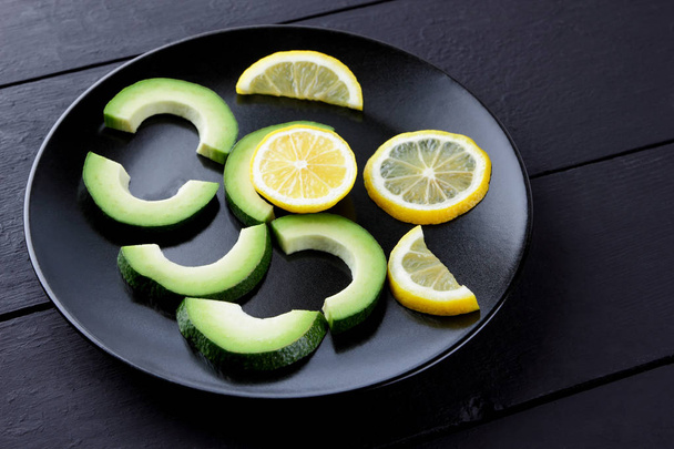 Avocado on a black plate top view, sliced avocado and lemon on black wooden boards, vegetarian food, useful natural food, tropical fruits for breakfast, Chilean food, copy space - Photo, Image