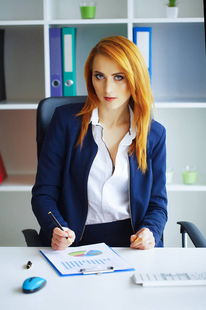 Üzleti. Egy komoly nő portréja. Üzleti vörös hajú nő ül a pultnál a nagy fény hivatalban egy mappát a kezét, és azt írja. Fehér inget és fekete szoknya lánynak. - Fotó, kép