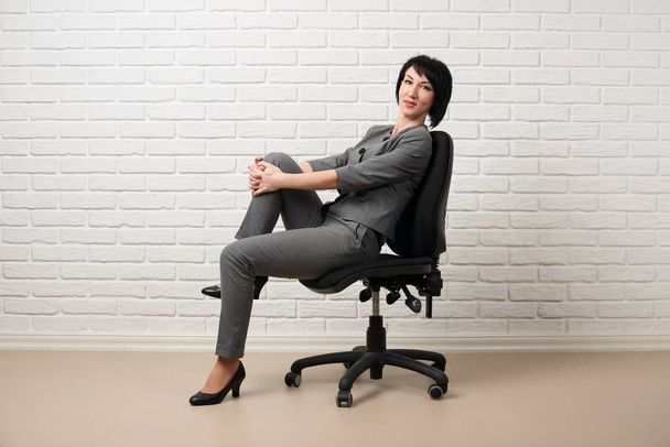 de zakenvrouw zittend op een stoel, gekleed in een grijs pak houdingen in de voorkant van een witte muur - Foto, afbeelding