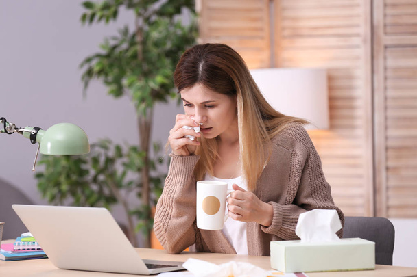 Sad exhausted woman with tissue and cup of hot drink suffering from cold while working with laptop at table - Foto, Bild
