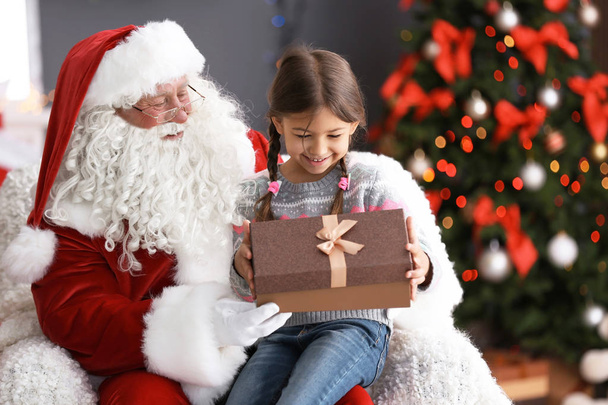 Little girl with gift box sitting on authentic Santa Claus' lap indoors - Фото, изображение