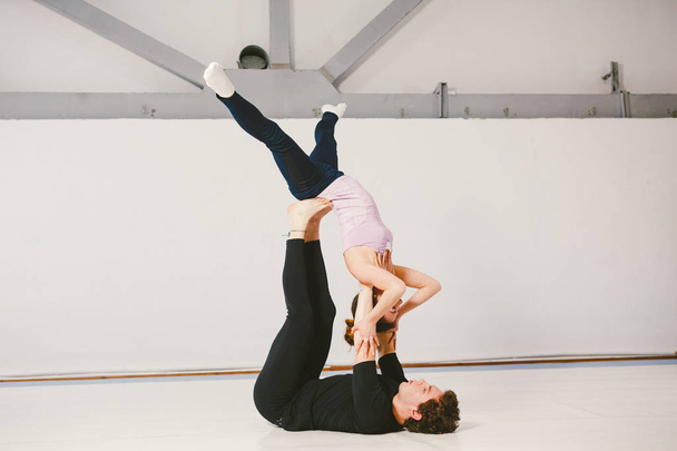 Молодая кавказская пара практикует акробатическую йогу в белом зале на ковриках. Они в позе Звезды.
. - Фото, изображение