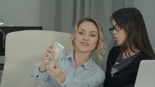 Two female colleagues taking selfies with phone sitting at the desk - Foto, Imagem