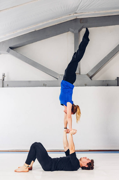Theme is sports and acreage. A young Caucasian male and female couple practicing acrobatic yoga in a white gym on mats. a man lies on his back and holds a woman on extended arms. Pose HAND-TO-HAND. - Photo, Image