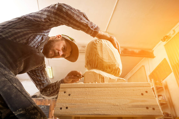 Jovem carpinteiro em uma roupa de trabalho serra a cabeça de um homem com uma árvore, usando um cinzel na oficina, em torno de um monte de ferramentas para o trabalho
 - Foto, Imagem