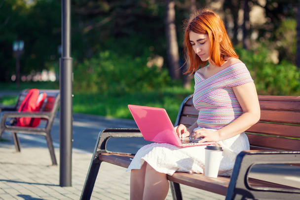 A young red-haired woman in a white romantic skirt, a pink top and blue sneakers running on a laptop and sitting on a bench on a summer day on in the city park - Фото, зображення