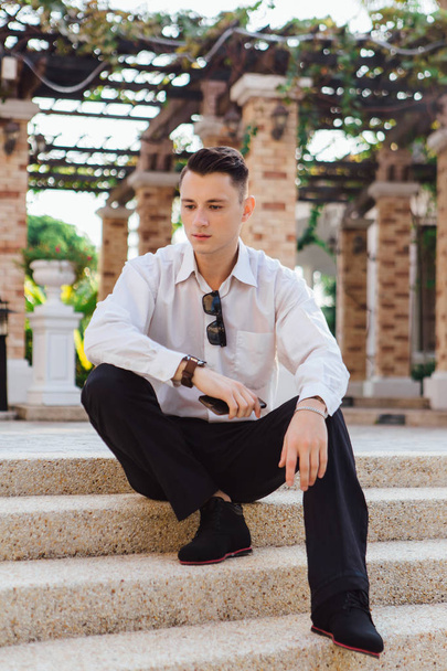 Young businessman holding his phone and sitting on stairs outdoors in a tropical country - Fotografie, Obrázek