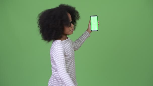 Young cute African girl with Afro hair showing phone and giving thumbs up - Imágenes, Vídeo