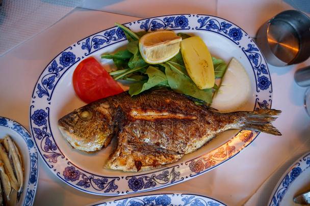 Tuore grillattu kokonaisen meriahvenen kala paahdetulla perunalla, vihreällä salaatilla, tomaatilla ja keltaisella limetillä valkoisella soikealla levyllä, jossa sininen kuvio Galatan sillalla, Istanbulissa, Turkissa
 - Valokuva, kuva