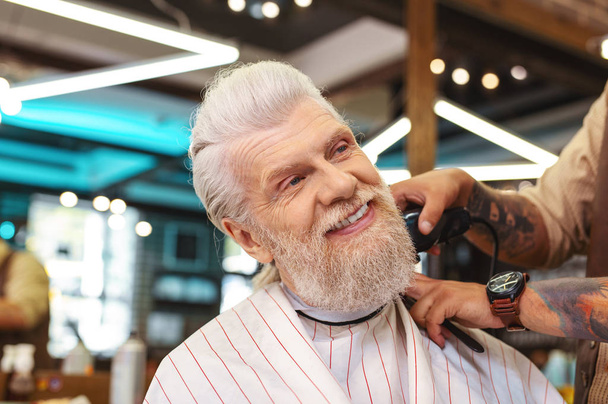 Pleased atmosphere. Delighted bearded senior man keeping smile on his face while vising barbershop - Photo, Image