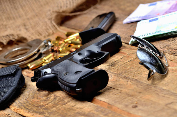 Pistolet 9mm - pistolet, menottes, balles, lunettes de soleil, billets en euros, chargeur et étui sur une vieille table en bois
 - Photo, image