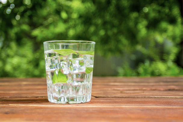 Um copo com uma bebida refrescante fria com gelo e folhas de hortelã em uma mesa de madeira e no fundo do Jardim. Conceito de recreação ao ar livre
. - Foto, Imagem