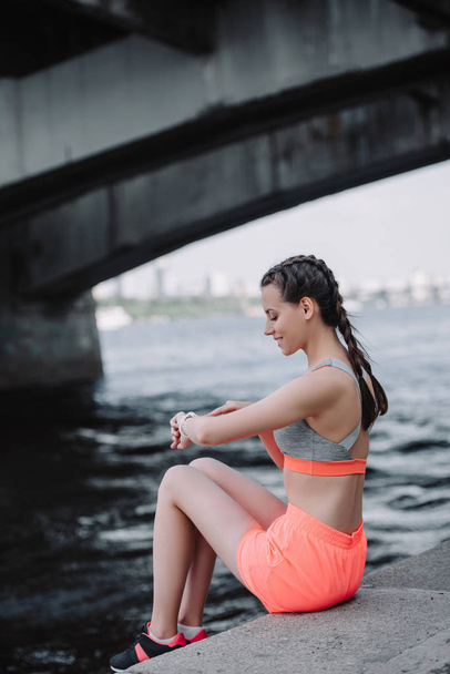 sportswoman looking at smart watch and sitting on quay - Foto, Bild