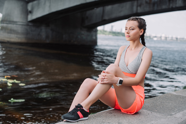 attractive sportswoman in sneakers sitting on quay near river - Foto, Bild