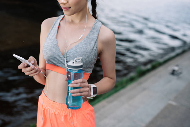 vue recadrée de la jeune sportive tenant smartphone avec écouteurs et bouteille de sport avec de l'eau
 - Photo, image