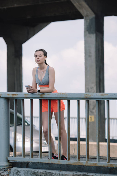 esportista pensativo com fones de ouvido e smartphone em pé perto de grades na ponte
 - Foto, Imagem