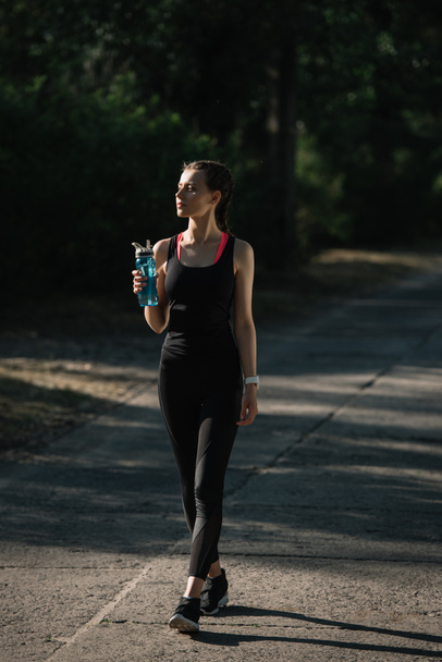 athletic woman holding sports bottle and walking on path in park - Foto, Imagen