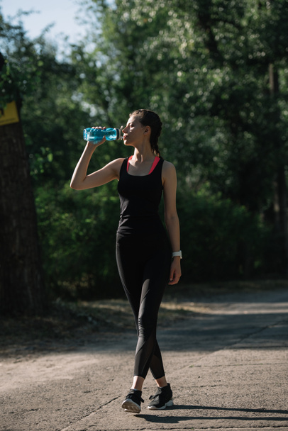 attractive athletic woman drinking water from sports bottle in park  - Photo, Image