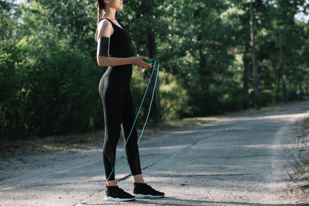 cropped view of sportswoman with smartphone in armband holding jump rope in park - 写真・画像