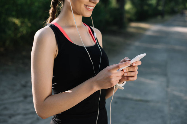 cropped view of smiling sportswoman listening music with earphones and using smartphone - Fotografie, Obrázek