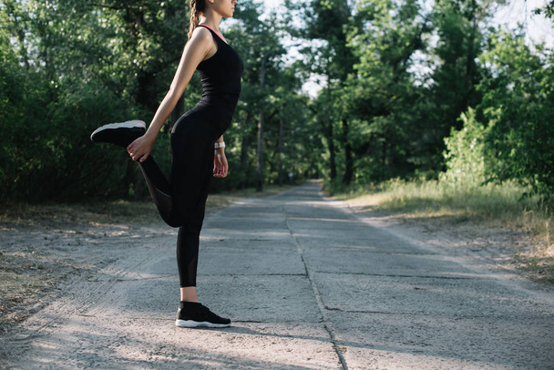 cropped view of sportswoman stretching leg on road in park  - Fotografie, Obrázek