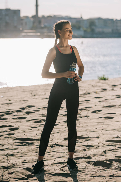 beautiful sportswoman with water in sports bottle standing on beach - Foto, Imagem