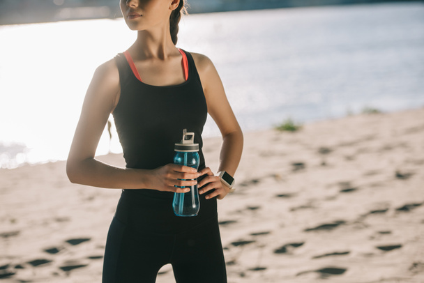 cropped view of athletic girl with sports bottle of water standing on beach - 写真・画像