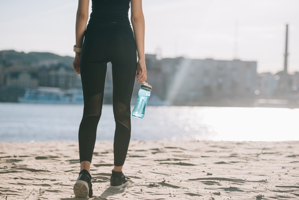 cropped view of sportswoman with water in bottle walking on beach with back light - Photo, Image