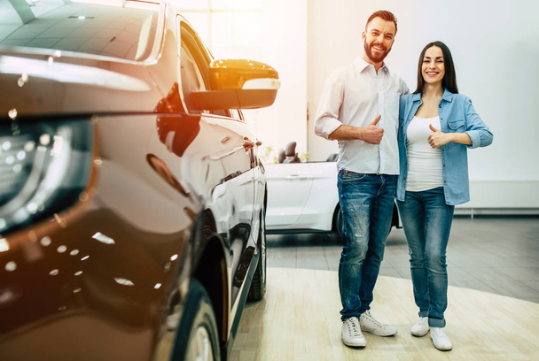 Happy young couple in casual wear standing near new car in dealership and showing thumbs up - Foto, immagini