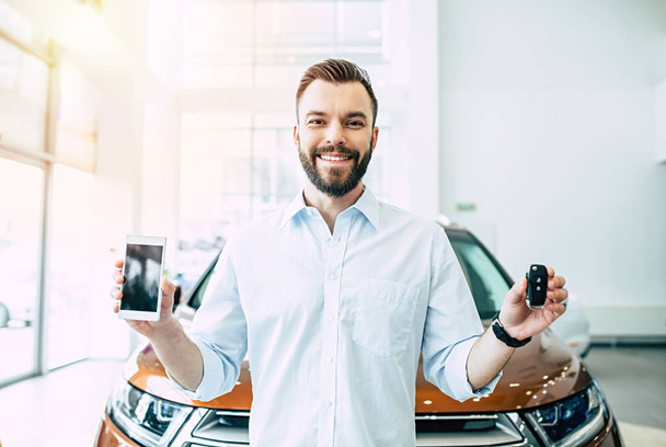 Happy handsome man standing with car key and smartphone on car background in dealership - Photo, Image