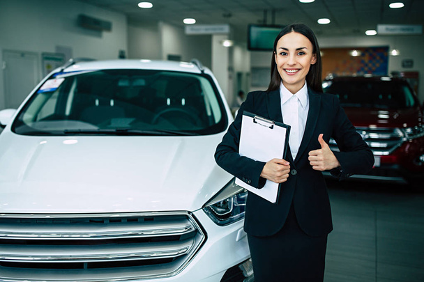portrait of cheerful smiling businesswoman in black suit holding documents and showing thumb up standing on dealership cars background - Photo, Image