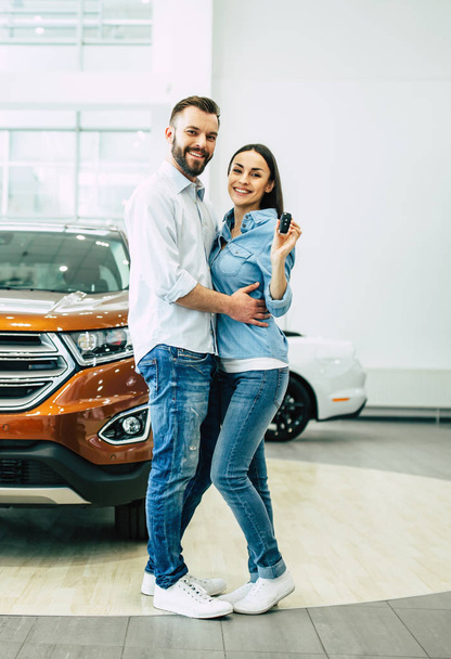Happy couple hugging and showing key standing on car background in dealership, full length - Фото, зображення