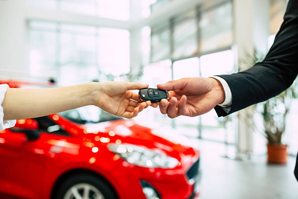 woman receiving key of new car from salesman in dealership, close up of hands on background of modern red car - Fotó, kép