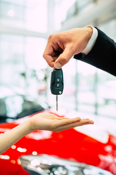 woman receiving key of new car from salesman in dealership, close up of hands on background of modern red car - Foto, afbeelding