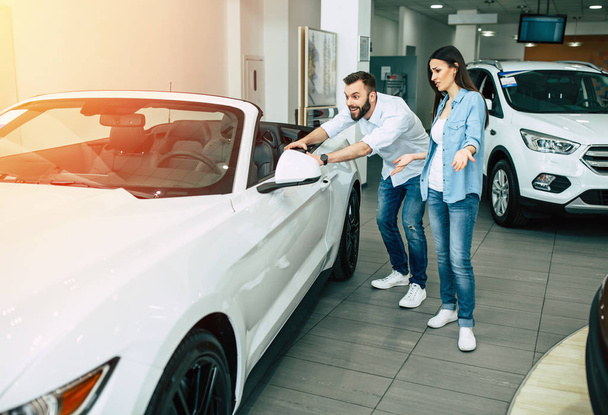 Happy young couple choosing new car in dealership, exciting man touching car - 写真・画像
