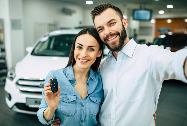 Happy young couple in casual wear standing near new car in dealership and showing key - Fotoğraf, Görsel