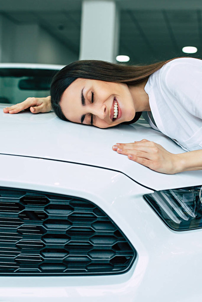 happy beautiful girl hugging hood of new car in dealership - Zdjęcie, obraz