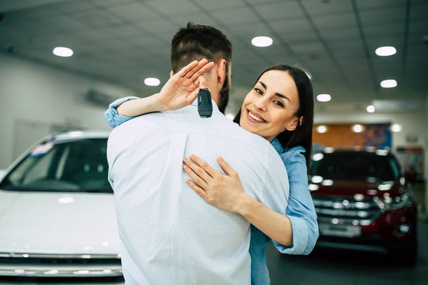 Happy young couple hugging standing in dealership, woman showing car key  - Photo, image