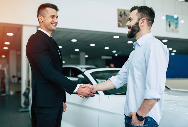 Two handsome businessmen shaking hands standing on background of dealership cars - Zdjęcie, obraz