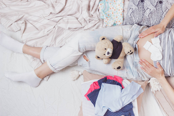 Mujer embarazada con un oso de peluche y varias ropas para un recién nacido, vista superior
 - Foto, imagen