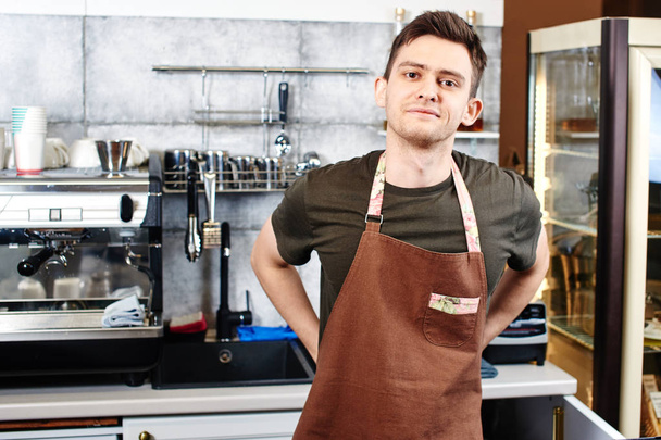handsome waiter in apron looking at camera while standing near coffee machine at workplace  - Foto, Imagem